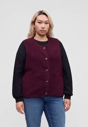 Ung person med blondt hår iført en knappet vinrød fleecevest over en sort langærmet skjorte og blå jeans, stående mod en ensfarvet baggrund.