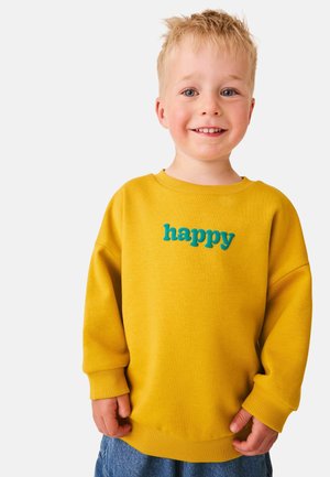 Młody blond chłopiec uśmiecha się, nosi żółtą bluzę z napisem „happy” niebieskimi literami i niebieskie dżinsy, stoi na jednolitym tle.