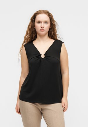 Kvinde med langt krøllet hår iført sort ærmeløs top med nøglehulskæring og beige bukser, stående mod en ensfarvet lys baggrund.