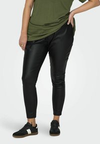 Zwarte nepleer broek met een tapered fit en elastische tailleband, gecombineerd met een losse olijfgroene top en zwarte sneakers met witte accenten.