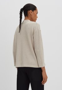 Lichtbeige geribbelde gebreide trui met een ontspannen pasvorm, wijde mouwen en een ronde halslijn, gecombineerd met zwarte broeken, gezien van achteren.