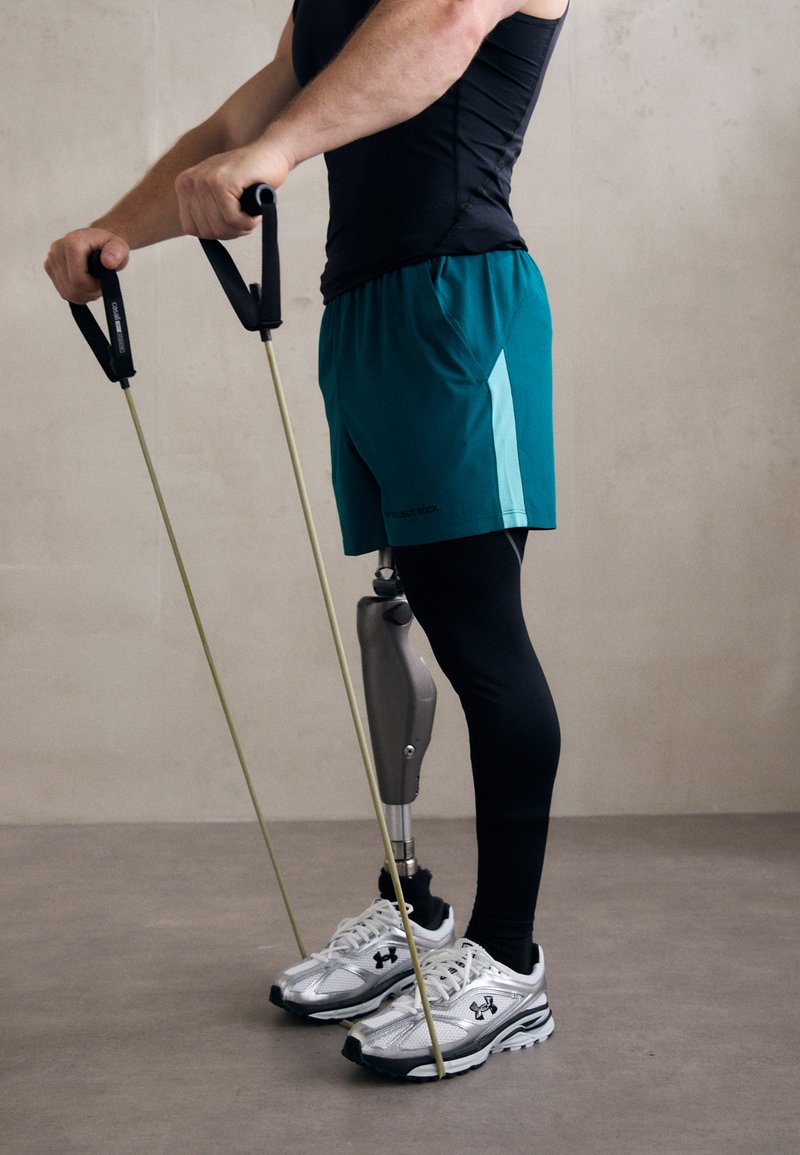 Athletic gear includes a black tank top, teal shorts, and black leggings. A silver sports shoe is worn on one foot, and a prosthetic leg is visible.