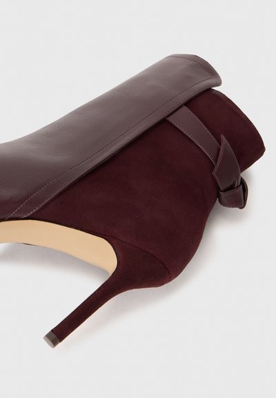 Zapato de tacón alto burdeos con parte superior de cuero liso, cuerpo de ante, puntera puntiaguda y detalle decorativo de correa y lazo en el tobillo.