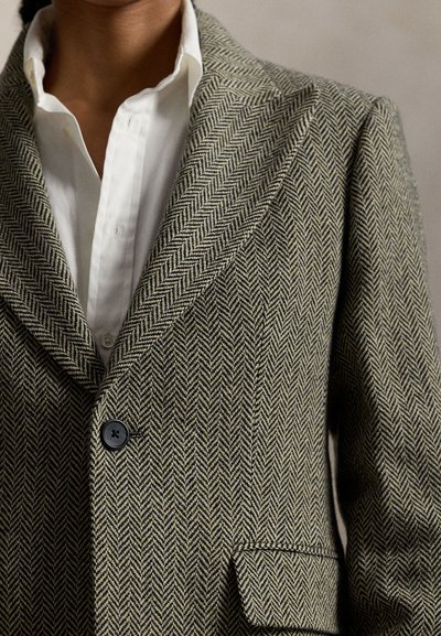 Gros plan d'une personne portant un blazer gris à motif chevrons sur une chemise blanche à col, montrant les détails des boutons et du revers.