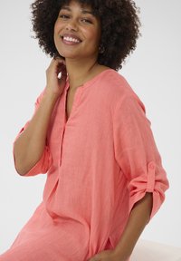 Camicia di lino corallo con vestibilità rilassata, scollo a V e maniche arrotolate. Tessuto testurizzato con leggere pieghe e spacchi laterali per una migliore circolazione dell'aria.