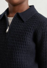 Tamno plavi pleteni pulover s patentnim zatvaračem, okovratnikom i teksturiranim rešetkastim uzorkom; mekana vuna, s uskim fitom oko vrata i ramena.