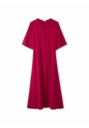 Donna in piedi con un lungo vestito rosso scuro con maniche larghe e cintura con nappine, indossa scarpe beige, su uno sfondo bianco semplice.