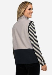 Gilet in pile grigio chiaro con colletto alto, abbinato a una maglietta a maniche lunghe a righe bianche e nere. Presenta un fondo di colore blu navy.