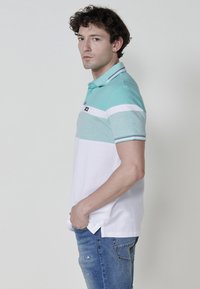 Menta zöld és fehér színblokkal díszített pólóing, rövid ujjakkal és gallérral, amelyen két gomb található.