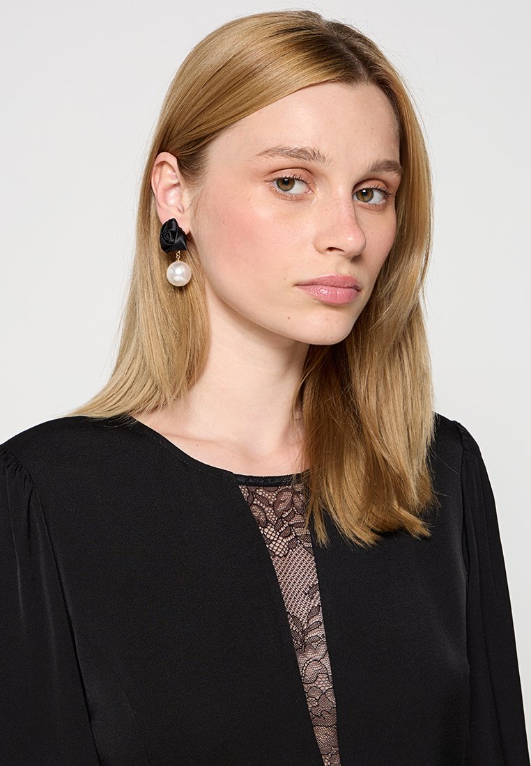 Vestido negro con panel de encaje, presenta pendientes redondos con acentos en negro y una gran perla, la modelo tiene el cabello rubio liso y maquillaje neutro.