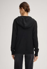 Zwarte gebreide hoodie met ribbeltextuur, losse pasvorm en lange mouwen. Heeft een hoge, ronde capuchon. Draagt met donkere denim broek.