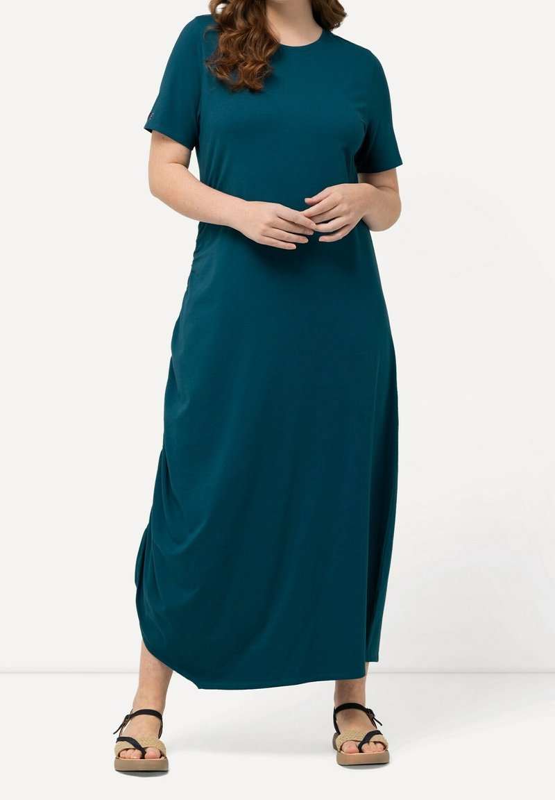 Femme portant une robe maxi à manches courtes couleur sarcelle avec un détail froncé sur le côté, debout avec les mains jointes et portant des sandales beige et noir.