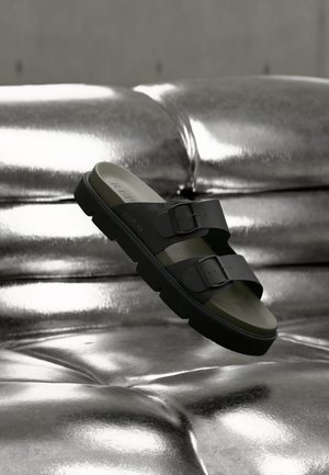 Schwarze Zwei-Riemen-Sandale mit Schnallen, Gummiaußensohle und einem olivgrünen Fußbett, oberhalb einer glänzenden Metalloberfläche positioniert.