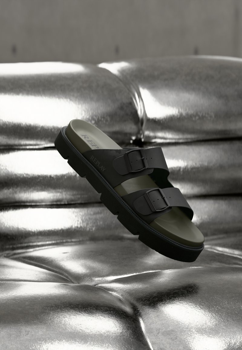 Μαύρο σανδάλι με δύο λουριά και αγκράφες, με καουτσούκ εξωτερική σόλα και φυτική πράσινη σόλα, τοποθετημένο πάνω σε γυαλιστερή μεταλλική επιφάνεια.