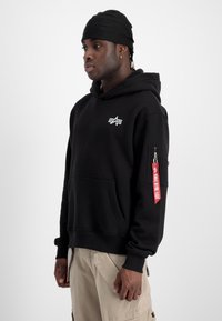 Schwarzer Hoodie aus Baumwolle mit einer vorderen Kängurutasche, seitlicher Zipp-Tasche und einem roten Etikett mit weißem Text an den Ärmeln.