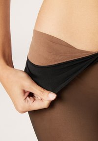 Opaka svarta och bruna strumpbyxor med en slät textur; en hand som drar ner den svarta midjebandet för att avslöja det bruna underlaget därunder.