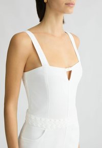 Combinaison blanche à bretelles larges, présentant un détail en forme de trou de serrure au niveau de la poitrine et une taille tressée. Tissu lisse et coupe ajustée.