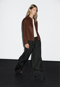 Giacca in misto lana marrone con colletto e bottoni, abbinata a pantaloni a gamba larga neri a righe. Il modello è in piedi contro uno sfondo semplice.