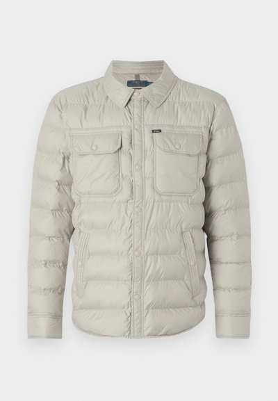 Polstret beige jakke med knaplukning fortil, to brystlommer og krave, der kan foldes ned. Har et vandret kviltet mønster og en glat tekstur.