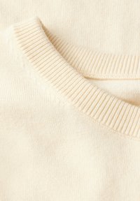 Primo piano di un maglione color crema, caratterizzato da un colletto girocollo a coste e da un tessuto morbido e strutturato con sottili motivi a maglia.