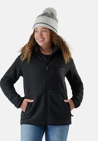 Black fleece jacket with front zip, side pockets, and a black nylon chest pocket. Model wearing a patterned knit hat and casual jeans.
