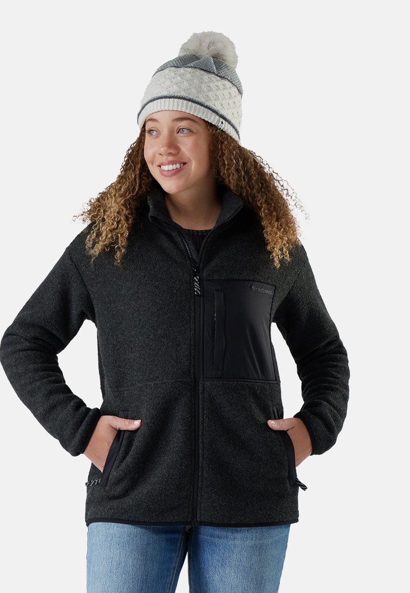 Black fleece jacket with front zip, side pockets, and a black nylon chest pocket. Model wearing a patterned knit hat and casual jeans.