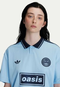 Lichtblauwe polo met marineblauwe accenten, geborduurd logo op de linkerborst en vetgedrukte "oasis" tekst op de voorkant. Zachte stof met subtiele strepen.