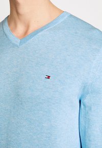 Himmelblauer V-Ausschnitt-Pullover aus weichem, strukturiertem Stoff, mit einem kleinen Logo auf der Brust in Rot, Weiß und Schwarz.
