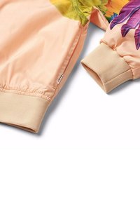 Ljus persikofärgad jacka med blommönster, ribbade ärmar och slät textur. Har en avslappnad passform och elastisk nederkant för komfort.