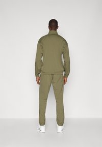 Ελαιόχρωμο αθλητικό σύνολο με ζακέτα με φερμουάρ και παντελόνι με ελαστική μέση; περιλαμβάνει γιακά σταντ και μανσέτες, συνδυασμένο με λευκά αθλητικά παπούτσια.