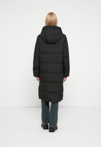 Inimene pikkade blondide juustega, kandes musta kapuutsiga sulejope, siniseid teksaseid ja musti klotsiga jalanõusid, seisab valge seina ees.