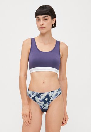 Lila Baumwoll-Sport-BH mit einem weißen elastischen Band, auf dem das Logo "Copenhagen Studios" zu sehen ist. Passender mehrfarbiger marmorierter Bikiniunterteil.