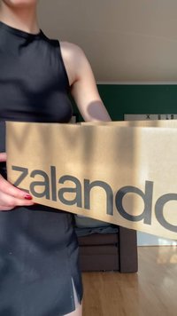 Caixa de cartão castanha com o logótipo preto "zalando" impresso em destaque de um dos lados, segurada por duas mãos contra um fundo suavemente iluminado.