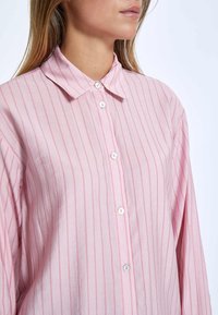 Camicia a righe rosa con colletto, chiusura a bottoni e tessuto leggero. Presenta righe verticali in morbide sfumature di rosa e rosso.