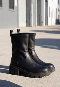 Paire de bottes en cuir noir mi-mollet avec semelles épaisses et languettes, posées sur un trottoir en béton ensoleillé près d’un bâtiment.