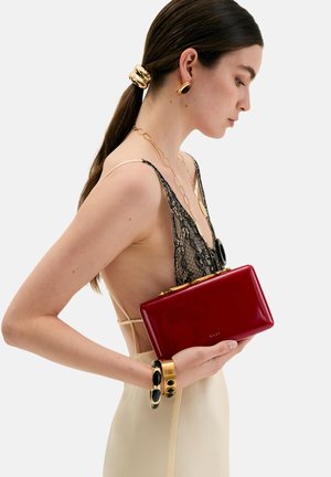 Žena s tmavými vlasy staženými do nízkého copu má na sobě béžové šaty s černou krajkou, zlaté a černé šperky a drží lesklou červenou kabelku u boku.