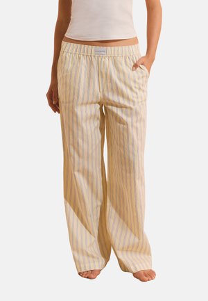 Donna che indossa pantaloni del pigiama beige larghi con sottili righe verticali e una cintura elastica, in piedi a piedi nudi con una mano in tasca.