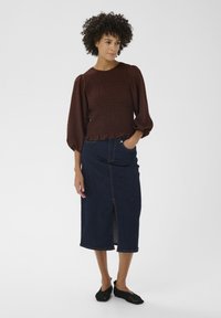 Haut smocké marron avec manches bouffantes, associé à une jupe midi en denim avec une fente à l'avant. Chaussures noires en mesh à enfiler complètent le look.