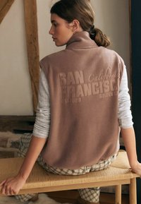 Brauner Fleece-Weste mit lässigen Armlöchern, mit geprägtem Text "SAN FRANCISCO" und "California 1907" auf dem Rücken. Getragen über einem grauen Oberteil.
