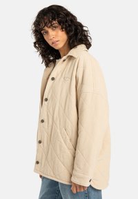 Beige quilted corduroy jacket with large button closure, side pockets, and a peaked collar. Features a small logo on the chest.