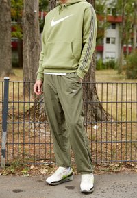 Olivgrön Nike-hoodie med vitt logga, matchande olivfärgade byxor med sidostråkar, och vita skor med gröna detaljer, mot en suddig parkbakgrund.