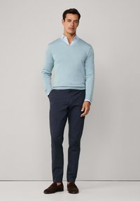 Svetlomodrý v-neck sveter preložený cez bielu košeľu s golierom, skombinovaný s tmavými námorníckymi nohavicami a hnedými semišovými topánkami. Jednoduchý, strihaný dizajn.