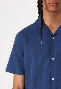 Short-sleeve shirt in dark blue fabric, button-up front, and a collar. Features a textured surface and subtle shine.