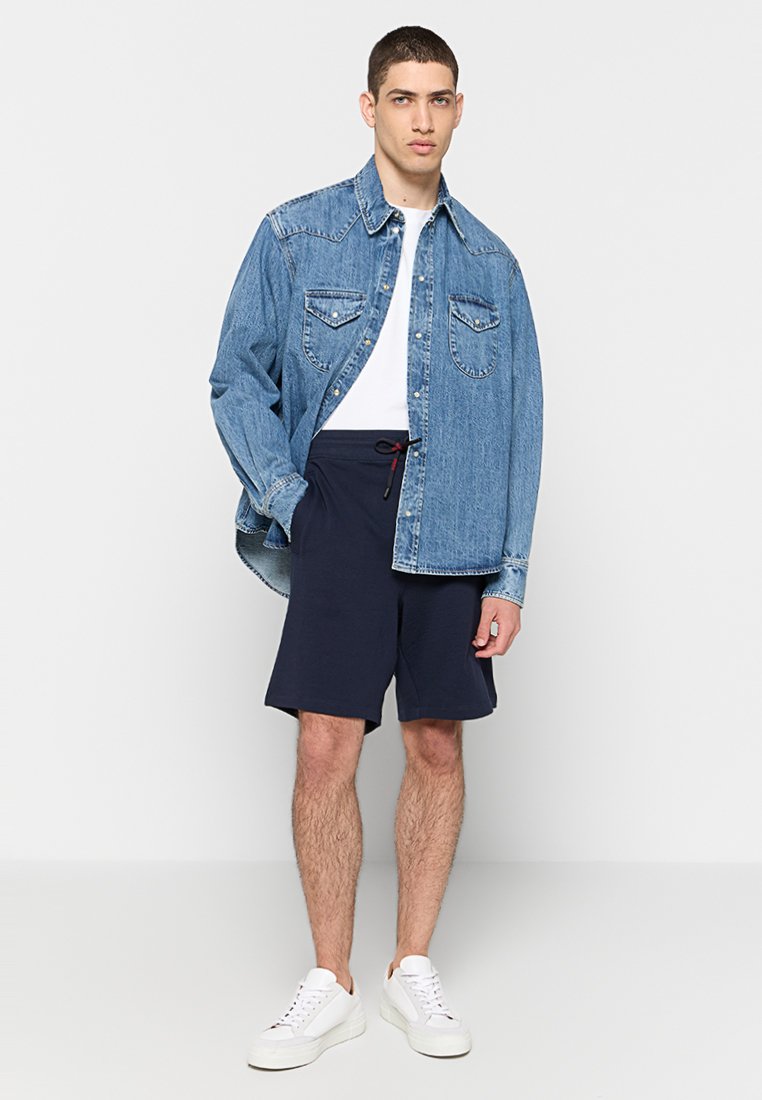 Denimhemd mit zwei Brusttaschen, weißem T-Shirt darunter, marineblauen Shorts und weißen Sneakers. Lässiger Schnitt, lange Ärmel und Button-Down-Design.