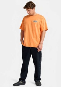 Oranžinė medvilninė trumpomis rankovėmis marškinėliai su juodu logotipu ant krūtinės, derinami su tamsiomis kelnėmis, stovint neutralioje fone.