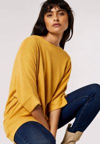 Pull surdimensionné jaune avec un col rond et des manches retroussées, associé à un jean bleu foncé et des bottines beige. Texture lisse.