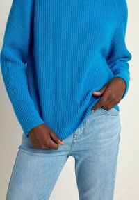 Hellblau gestrickter Pullover mit gerippter Textur, weiten Ärmeln und lockerer Passform, kombiniert mit hellblauen Jeans.