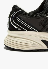 Baskets noires de sport avec une tige en mesh texturé, accents blancs et une semelle crème présentant un motif ondulé et des éléments de design courbés.