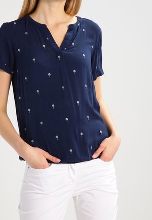 Femme portant un chemisier bleu marine à manches courtes avec de petits motifs blancs de palmiers et un pantalon blanc, mains dans les poches.