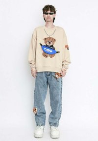 Sudadera beige con un gráfico de oso de peluche, riñonera azul, oso bordado en los jeans y zapatillas blancas gruesas en un atuendo casual.